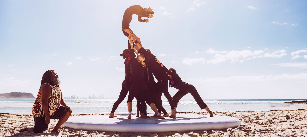 A group of acrobats on a beach, dressed in black, holding up one acrobat with a man kneeling to the side, drenched in beautiful sunlight