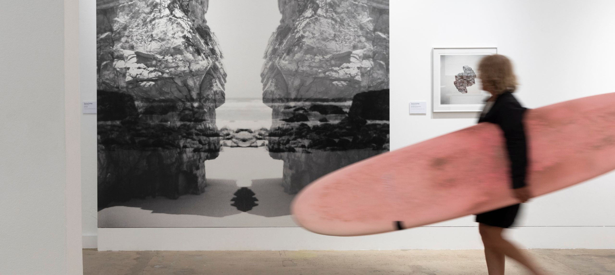 Man with pink surfboard walking through gallery looking at large black and white rock photography artwork