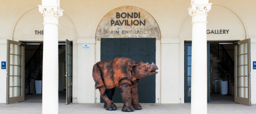 A giant puppet rhino standing between the Bondi Pavilion pillons with the ocean behind it