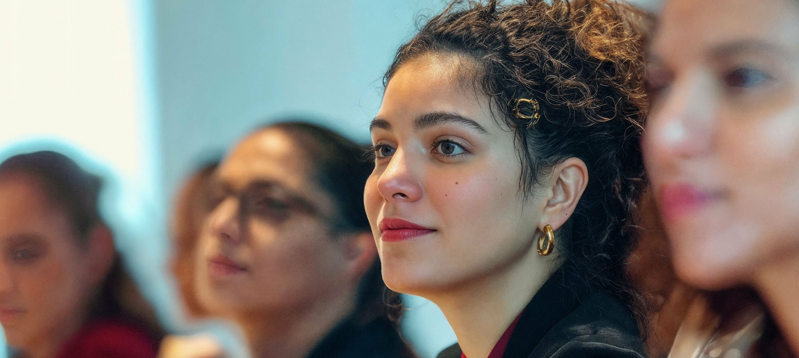 Women with brown curly hair and red lipstick looking ahead with other women blurred in background