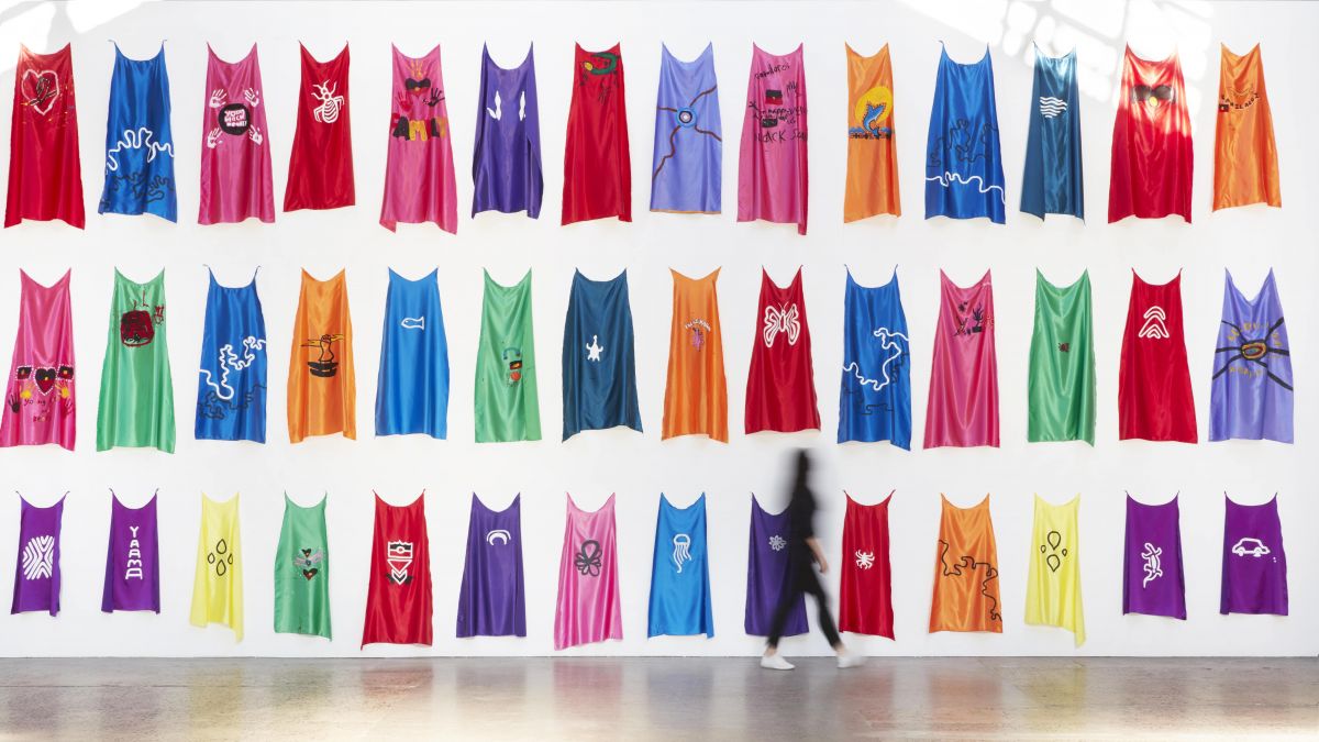 A large white wall with brightly coloured capes. The capes are three layers high and tower over a small visitor.