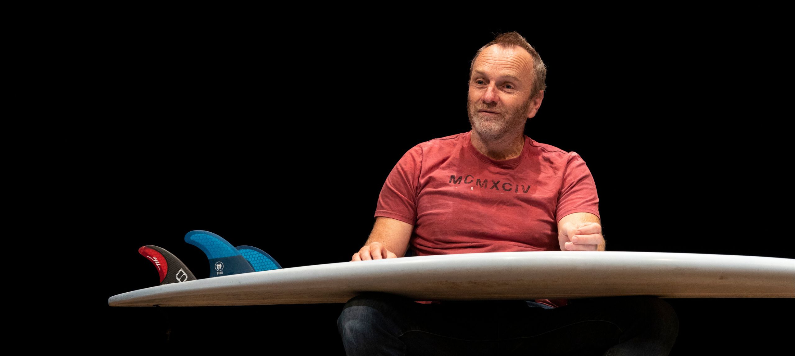 Man with red t-shirt sitting with a surfboard on his knee