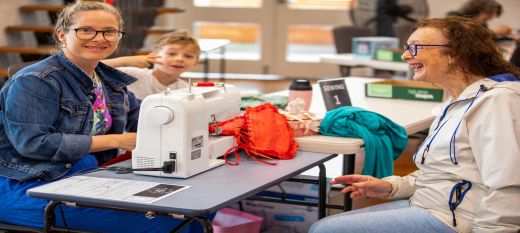 woman with sewing machine looking at camera and sitting across a lady laughing 