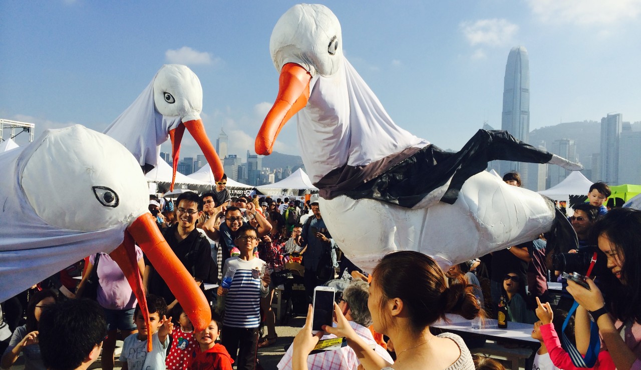 Three giant seagulls in a crowd. A woman takes a photo of the larger-than-life seagull puppets.