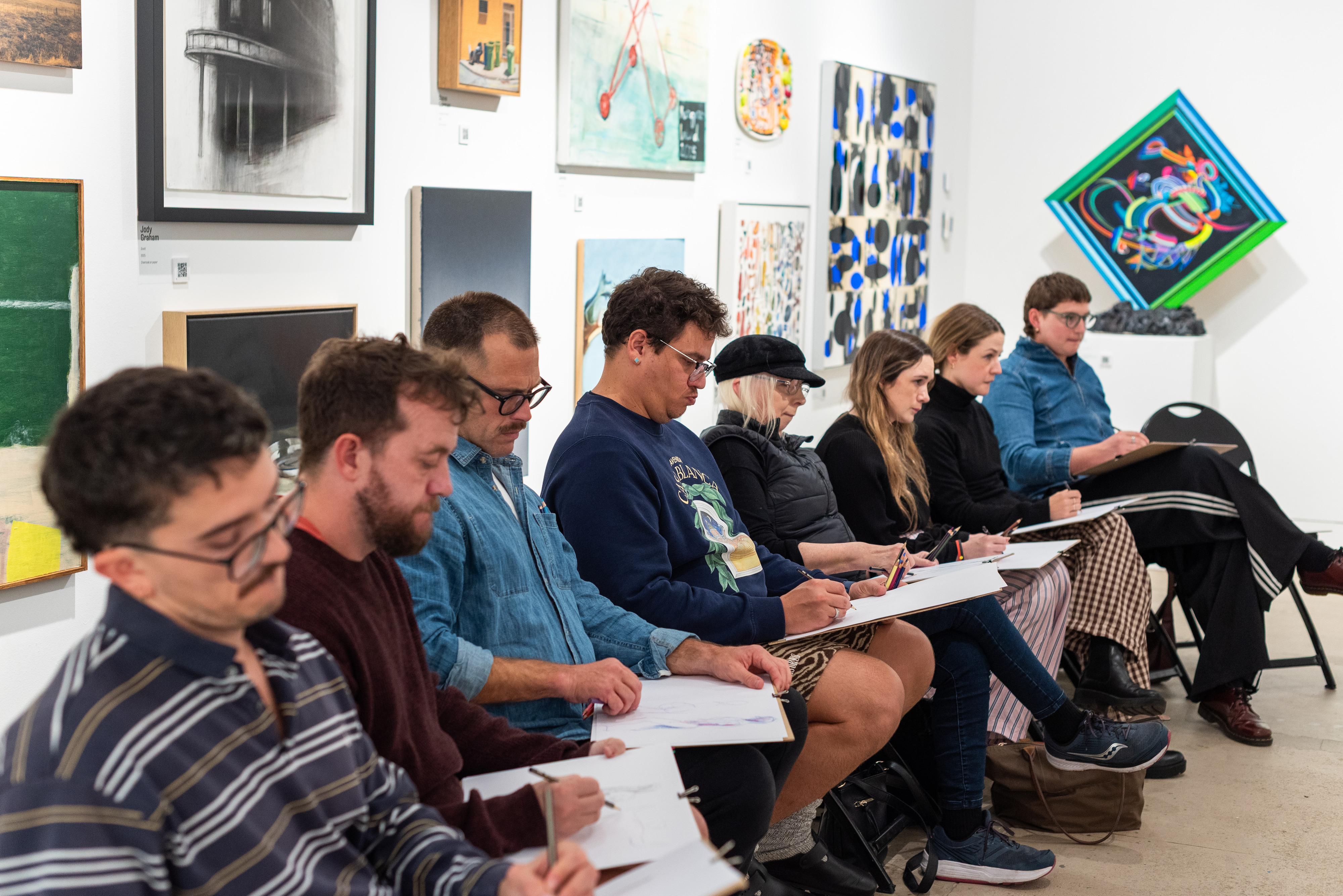 Line up of seated people working on drawing pads in a gallery