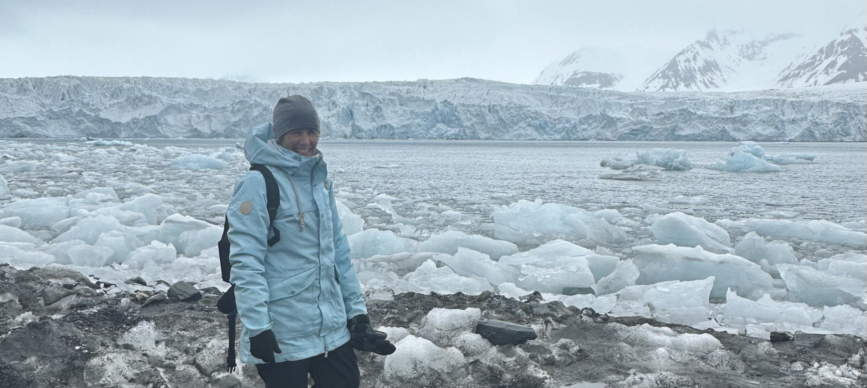 A photograph of Michele Barker in an Arctic landscape. She is wearing polar clothing, behind her is a white icy landscape with mountains in the distance. 
