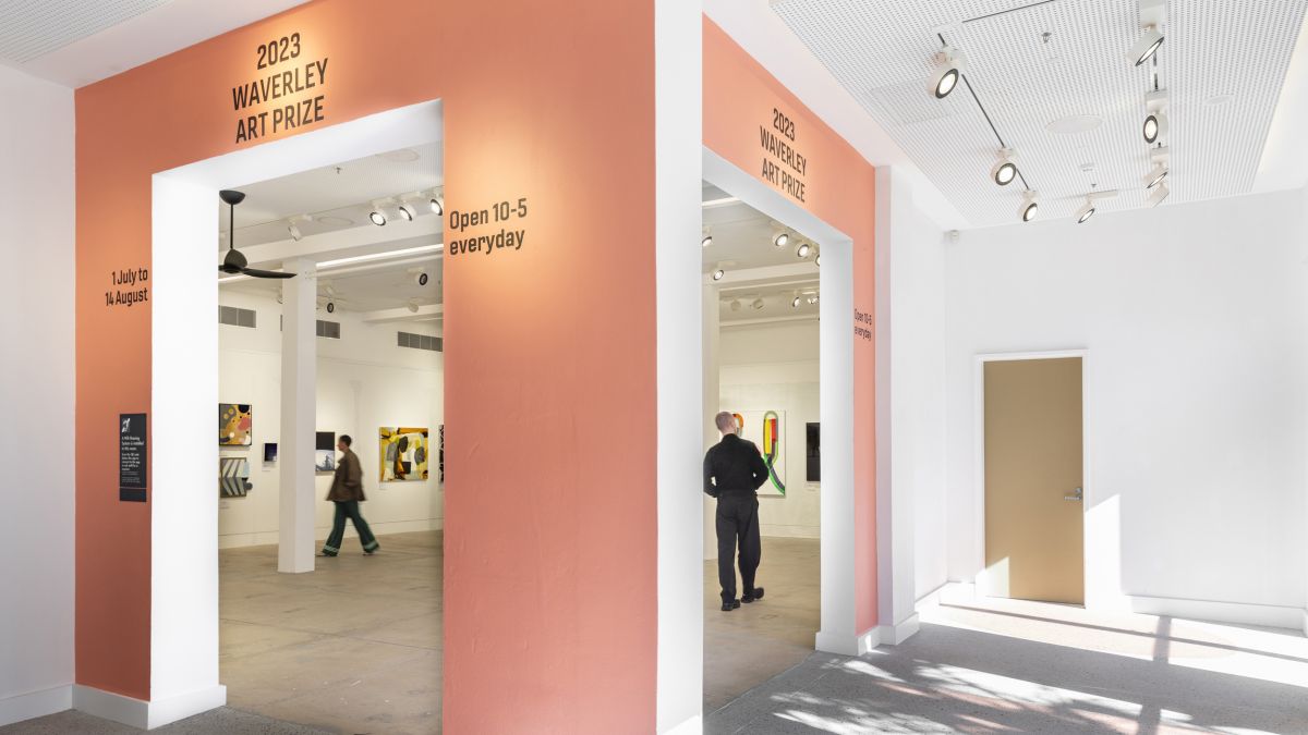 A photograph of the Bondi Pavilion Gallery entryway. It features a large orange opening with two doors, through it is a gallery full of works and 2 people. 