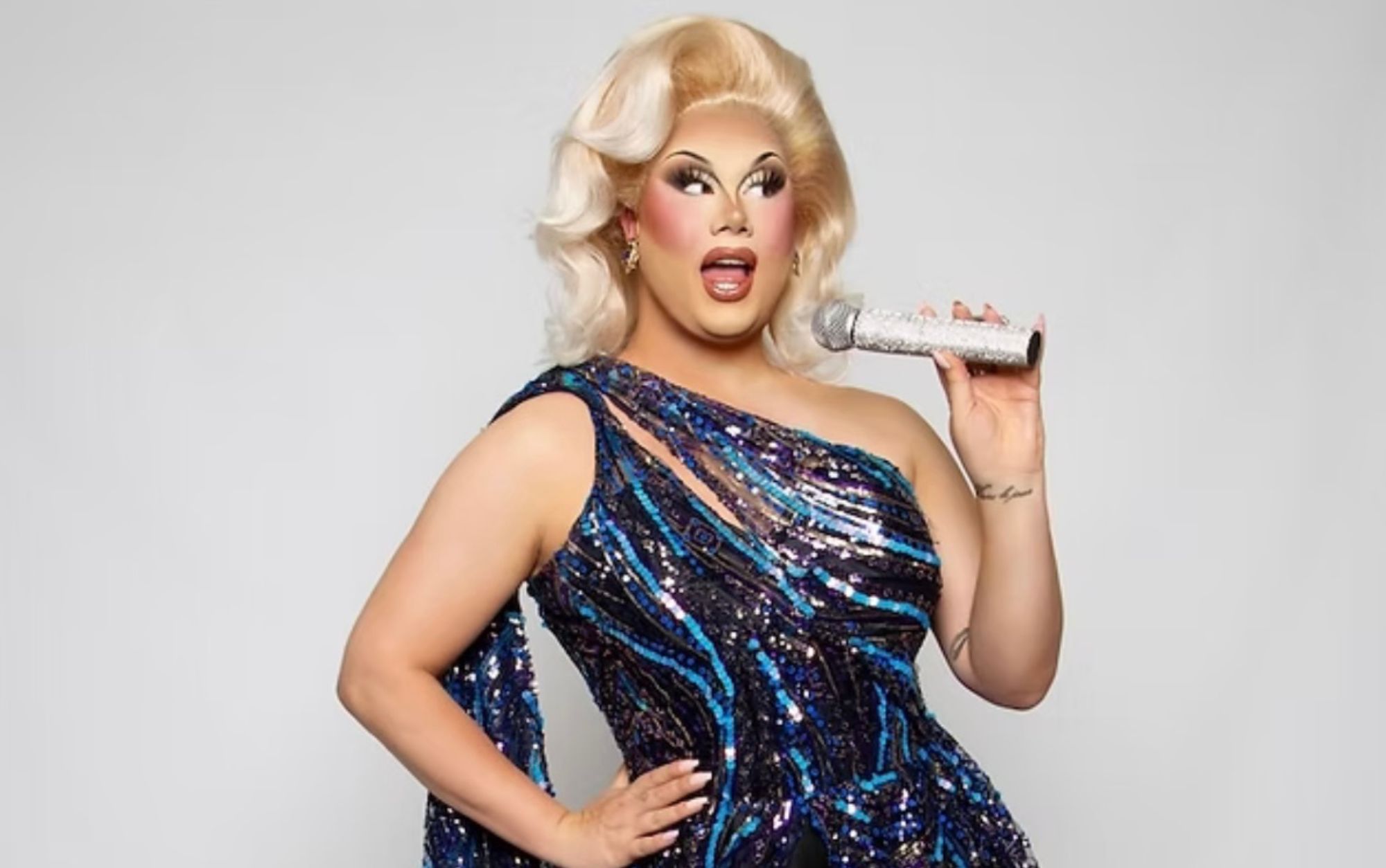 A drag queen, Vybe, with blonde hair and a sequined blue and black dress with a microphone held up in one hand and her other hand on her hip. Image courtesy of Untamed Entertainment