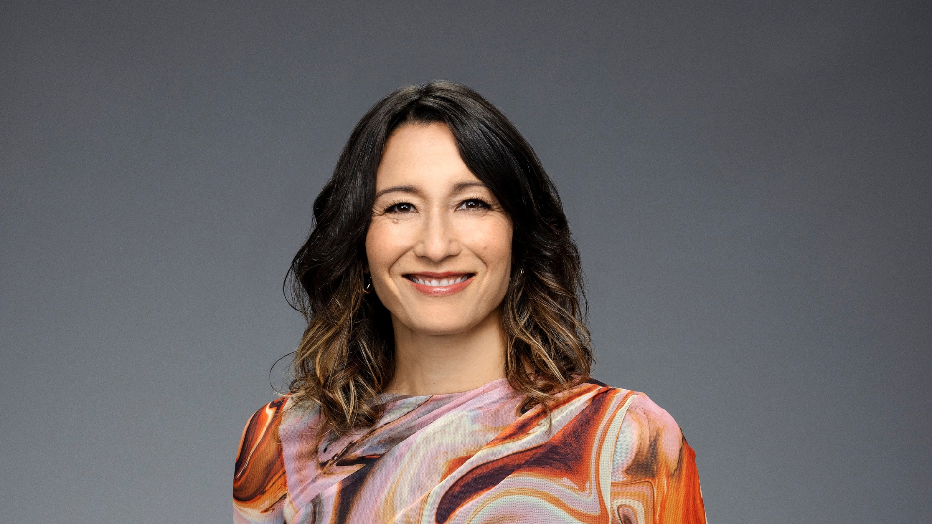 A woman with long dark hair and a brightly patterned top looks into the camera
