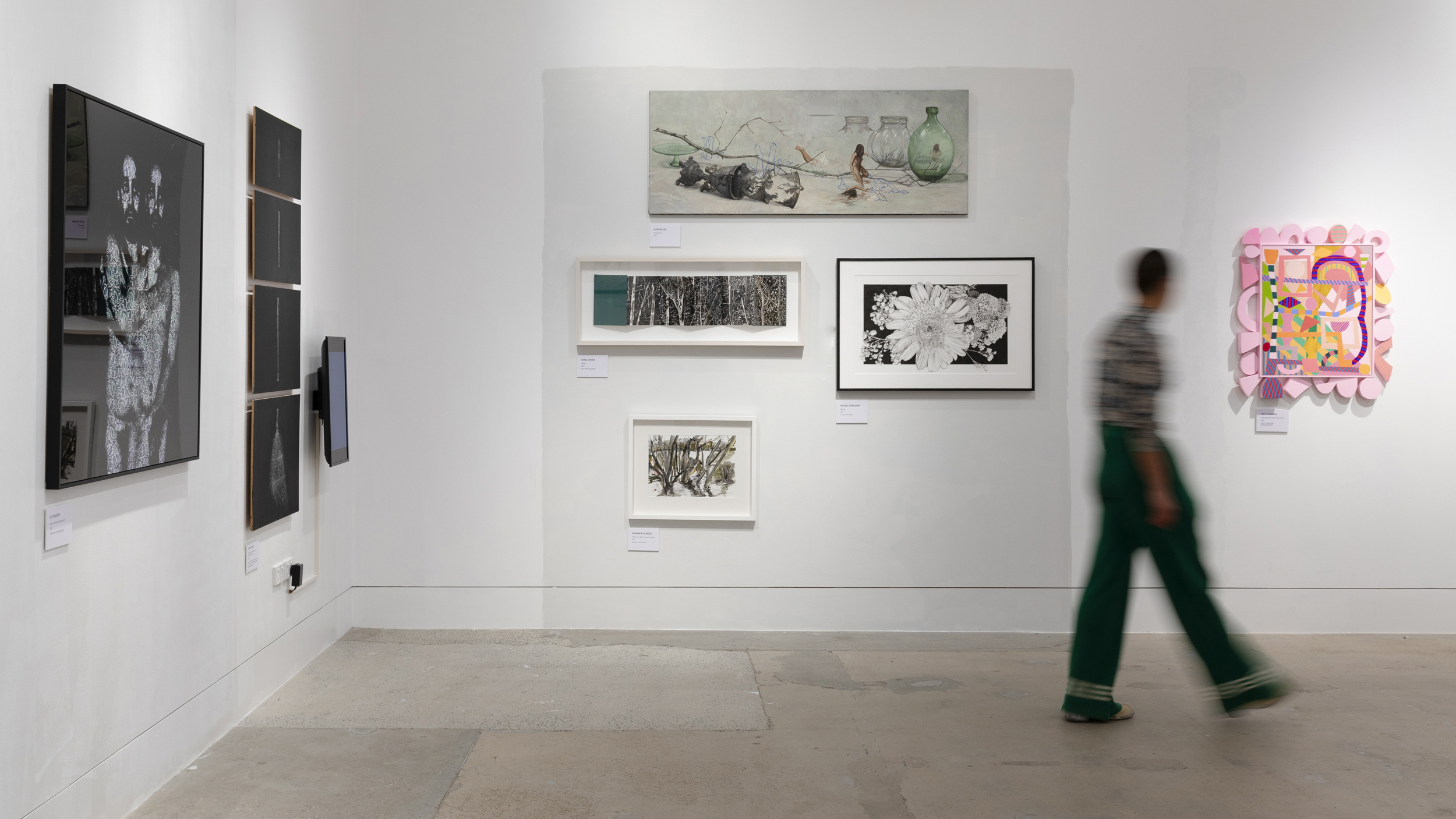 A photograph of a gallery corner, on the white walls are paintings and a screen. In front of it a blurry figure strides past.