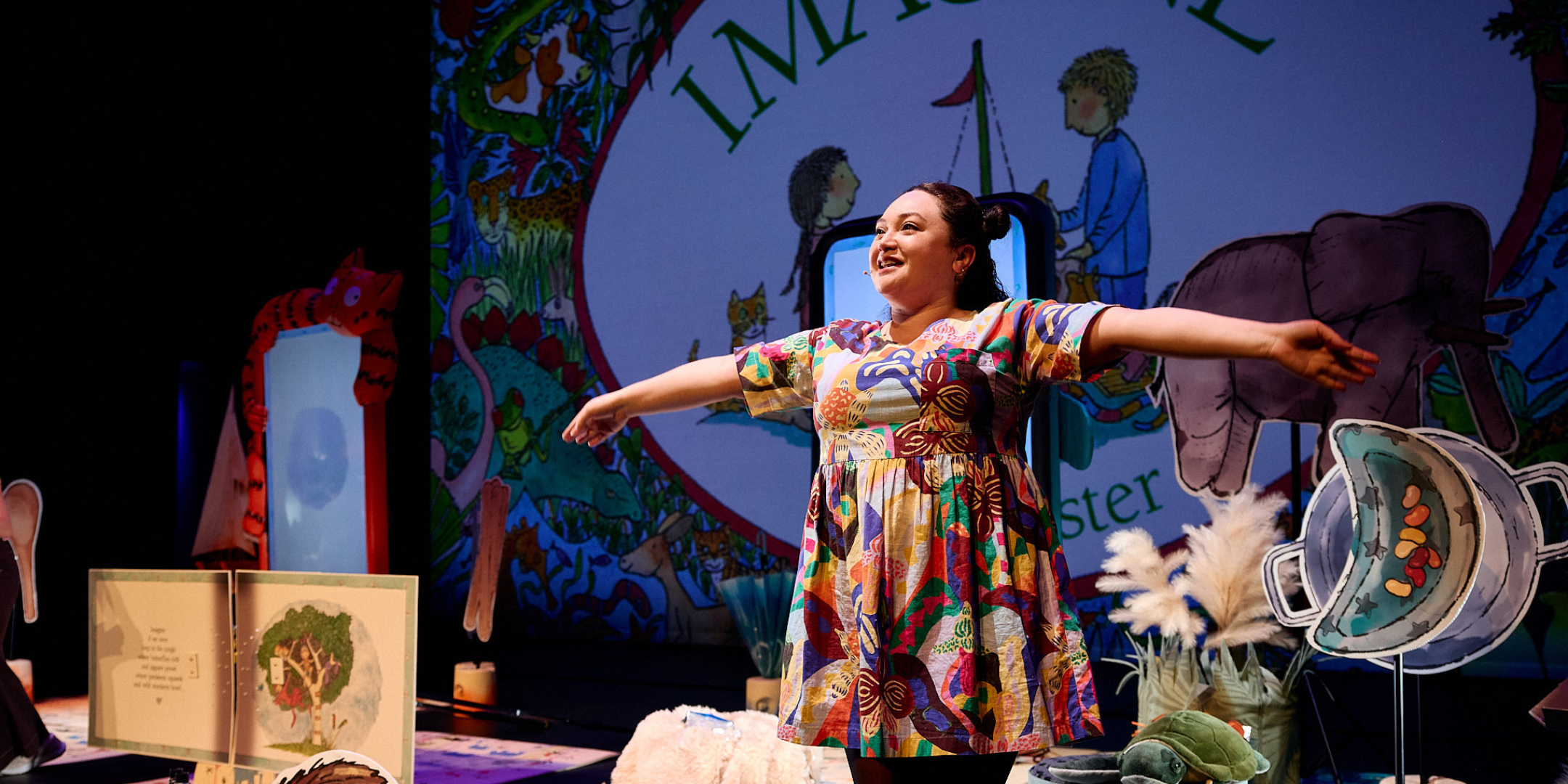 Girl in a colourful patterned dress singing on stage with arms outstretched surrounded by colourful objects