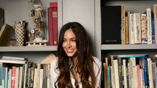 A woman with long flowing hair glances up in front of a large bookcase of books and treasures